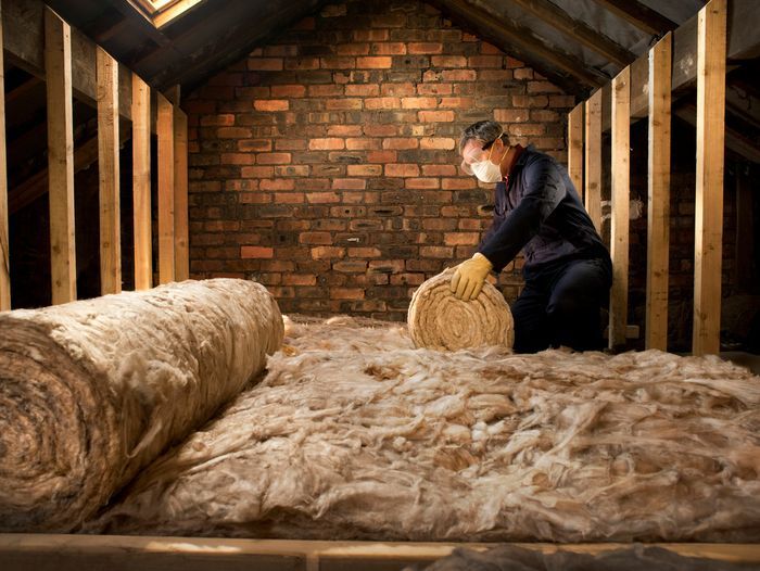 A worker in a protective mask and gloves installing rolls of fiberglass insulation, one of the common types of insulation, in an attic space.