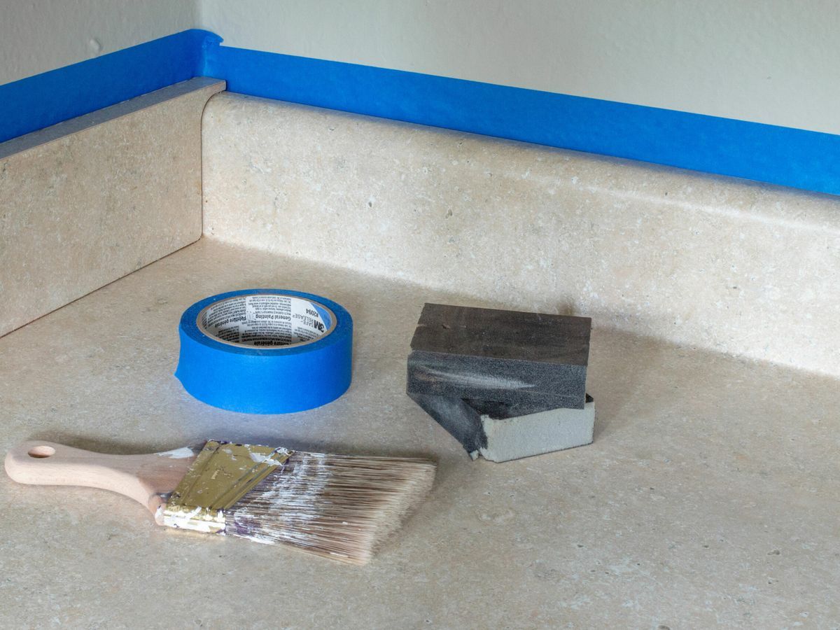 The supplies needed to prepare a laminate countertop, including a paint brush and blue tape.